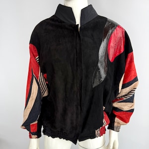 May include: Black suede jacket with a high collar and elastic waist. The sleeves feature red, black, and beige geometric patterns. The shoulder has a red and gray snakeskin-like design. A unique vintage jacket.
