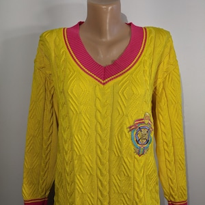 Pode incluir: Camisola de malha amarela com cabo, decote em V rosa e acabamento rosa em volta das mangas e da parte inferior. A camisola tem um logotipo bordado na frente com um tigre e o texto "D.C.F."