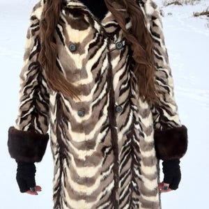 Può includere: Un lungo cappotto in ecopelliccia con un motivo a strisce zebrate nei toni del marrone, crema e nero. Il cappotto presenta una chiusura doppiopetto con bottoni scuri, un cappuccio e polsini in pelliccia marrone scuro. La persona indossa guanti neri.