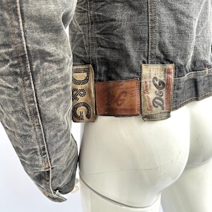 Puede incluir: Una chaqueta vaquera gris con un aspecto desgastado. La chaqueta tiene varios parches en la espalda, incluido uno que dice "D&G Denim".