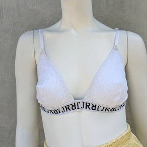 Puede incluir: Sujetador blanco de triángulo con un patrón de encaje blanco y una banda elástica negra con el texto "JRJRJRJRJRJRJRJR" repetido. El sujetador está en un maniquí blanco que lleva una falda de punto amarilla.