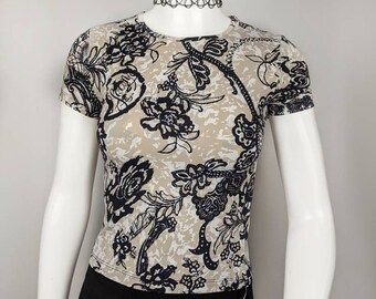 May include: A short-sleeved beige top with a black floral pattern. The shirt has a crew neck and is displayed on a mannequin. A black choker necklace is also visible.