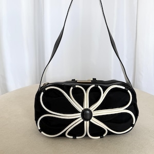 Puede incluir: Un bolso de hombro negro con una prominente aplicaci&oacute;n floral blanca en la parte delantera. El bolso tiene una correa larga negra, una forma rectangular redondeada y un acento circular negro central en el n&uacute;cleo de los p&eacute;talos.