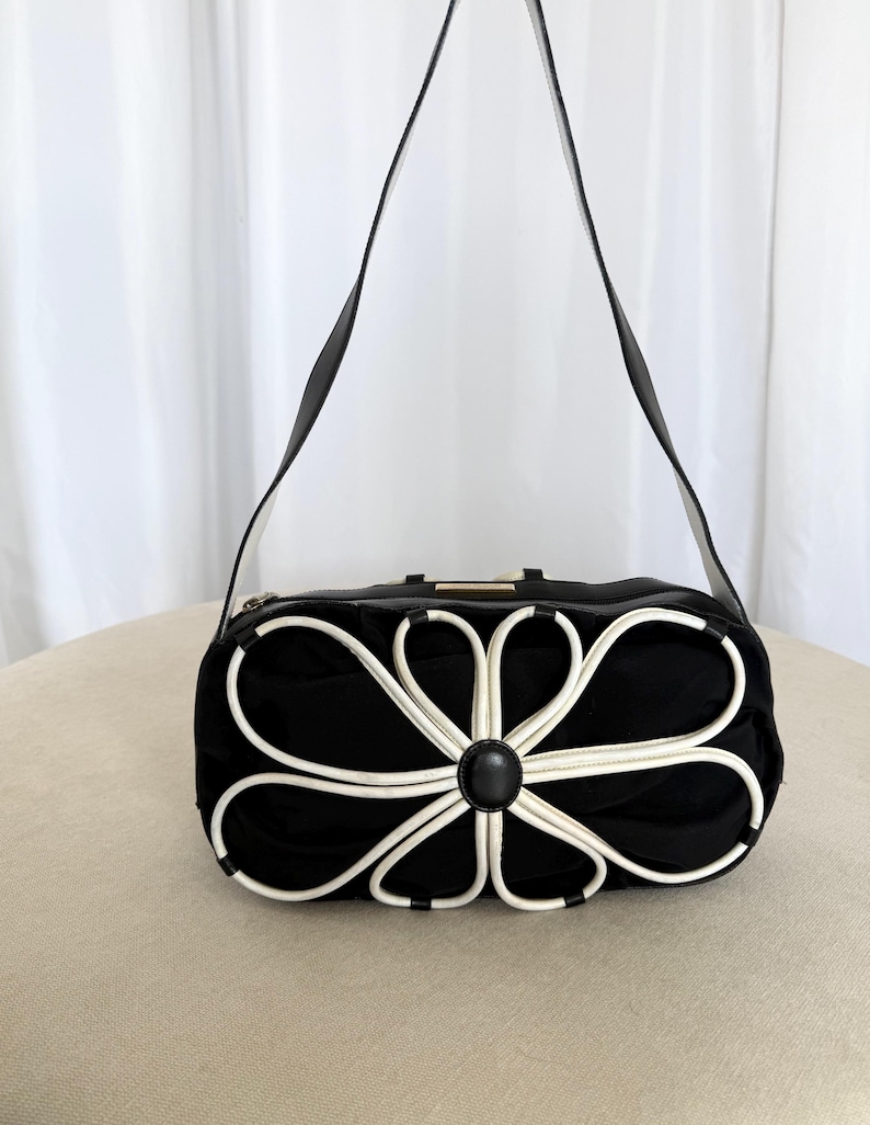 Puede incluir: Un bolso de hombro negro con una prominente aplicaci&oacute;n floral blanca en la parte delantera. El bolso tiene una correa larga negra, una forma rectangular redondeada y un acento circular negro central en el n&uacute;cleo de los p&eacute;talos.