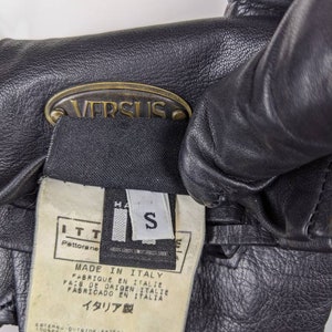 May include: Black leather gloves with a gold metal tag that says "VERSUS". The tag is attached to a black label with white text that says "MADE IN ITALY", "FABRIQUE EN ITALIE", "PAIS DE ORIGEN ITALIE", "FABRICADO EN ITALIA", and "イタリア製". The label also has a size tag that says "S".
