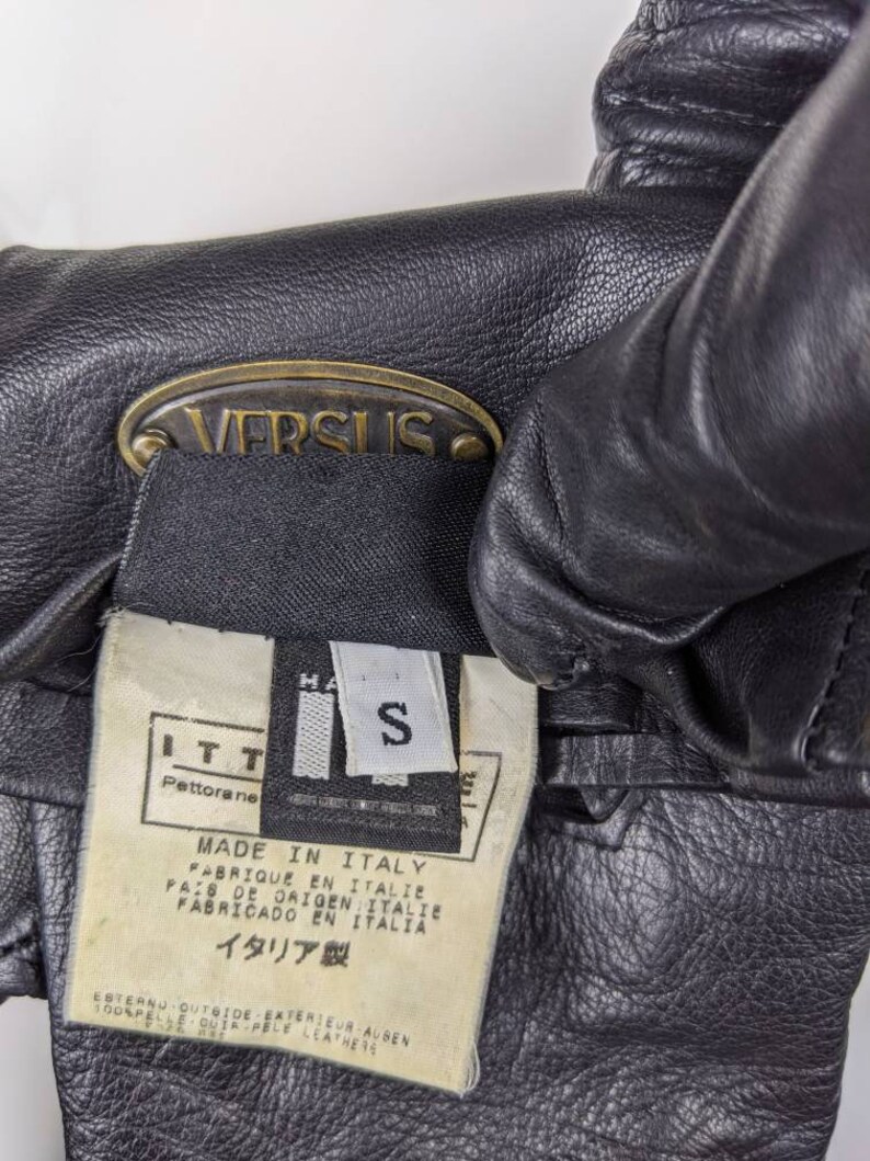 May include: Black leather gloves with a gold metal tag that says "VERSUS". The tag is attached to a black label with white text that says "MADE IN ITALY", "FABRIQUE EN ITALIE", "PAIS DE ORIGEN ITALIE", "FABRICADO EN ITALIA", and "イタリア製". The label also has a size tag that says "S".