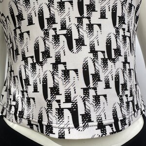 May include: White top with a black and white abstract print. The print features a repeating pattern of letters.