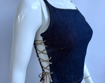 Puede incluir: Un top de mezclilla azul oscuro con tirantes finos y cuello cuadrado. Los lados presentan un diseño de cordones con lazos color beige y cuentas decorativas. El top se exhibe en un maniquí blanco.