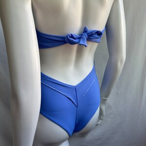 Pu&ograve; includere: Un costume da bagno blu pervinca con chiusura a fiocco sul retro. Il costume intero ha uno slip a taglio alto e un design a forma di V sul retro. Il costume da bagno &egrave; esposto su un manichino.