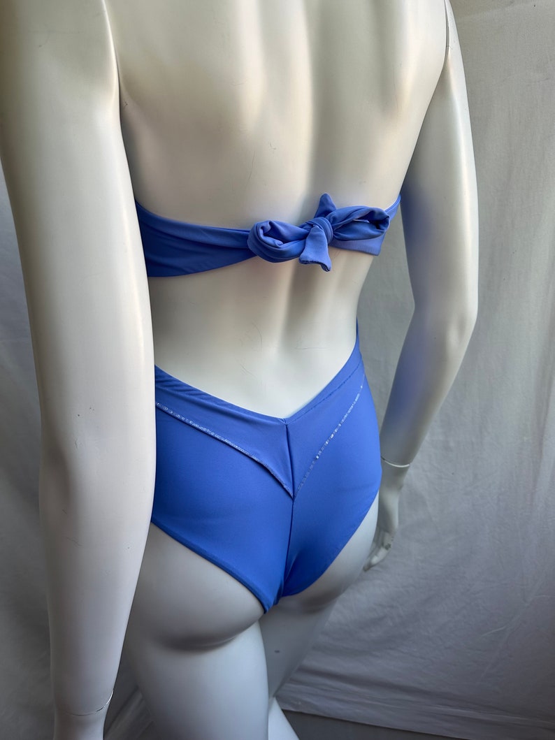 Pu&ograve; includere: Un costume da bagno blu pervinca con chiusura a fiocco sul retro. Il costume intero ha uno slip a taglio alto e un design a forma di V sul retro. Il costume da bagno &egrave; esposto su un manichino.