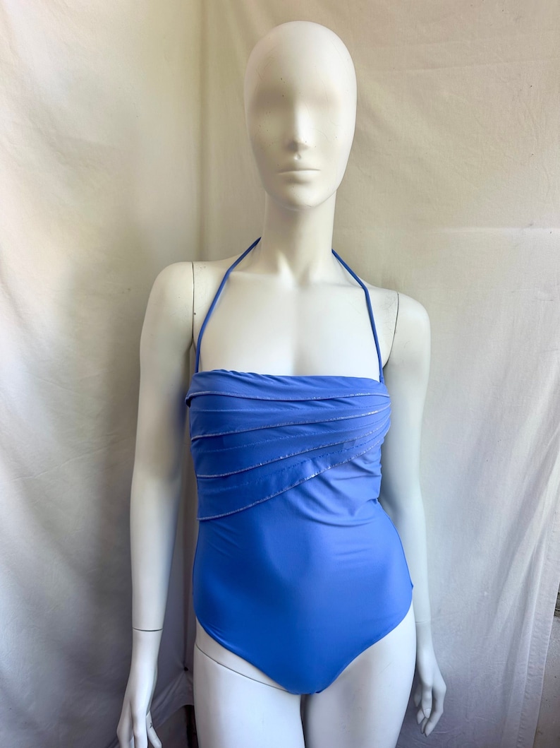 Pu&ograve; includere: Un costume da bagno blu pervinca con collo all'americana e corpetto arricciato. Il costume da bagno ha un top stile fascia con delicati accenti argentati. Il design intero &egrave; esposto su un manichino.