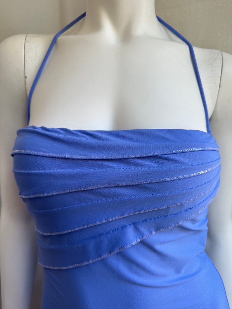 Pu&ograve; includere: Un abito a fascia blu pervinca con corpetto a strati. L'abito presenta un design senza spalline con cinturino all'americana ed &egrave; impreziosito da paillettes argentate lungo le cuciture a strati. L'abito &egrave; esposto su un manichino.