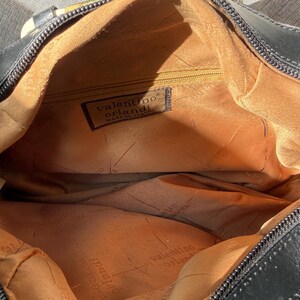 Puede incluir: Un bolso negro con forro interior color canela que presenta el logotipo y el texto de Valentino Orlandi. El interior incluye un bolsillo con cremallera y una etiqueta de tela que dice Valentino Orlandi Made in Italy. El bolso tiene cierre negro.