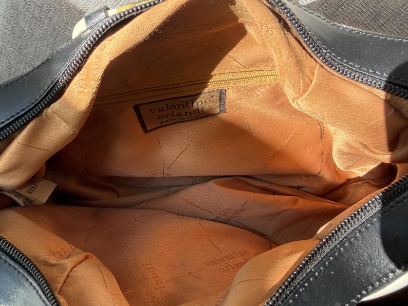 Puede incluir: Un bolso negro con forro interior color canela que presenta el logotipo y el texto de Valentino Orlandi. El interior incluye un bolsillo con cremallera y una etiqueta de tela que dice Valentino Orlandi Made in Italy. El bolso tiene cierre negro.