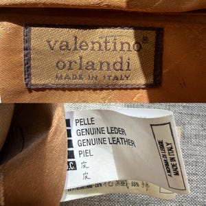 Puede incluir: Una etiqueta rectangular de tela marr&oacute;n con el texto bordado valentino orlandi MADE IN ITALY. Una etiqueta blanca de cuidado enumera materiales en varios idiomas, incluyendo PELLE, GENUINE LEATHER, PIEL, e incluye A NORMA DI LEGGE MADE IN ITALY.