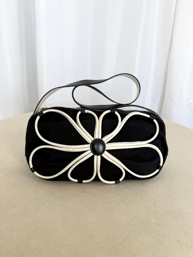 Puede incluir: Un bolso de mano negro con una llamativa aplicaci&oacute;n floral blanca en la parte delantera. El dise&ntilde;o consta de ocho bucles blancos en forma de p&eacute;talos que irradian desde un bot&oacute;n circular negro central. El bolso incluye una correa negra y cierre de cremallera.