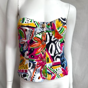 Pode incluir: Um top curto sem alças com uma estampa abstrata colorida. O top tem um fundo branco com formas e linhas abstratas de cor rosa brilhante, laranja, amarelo, verde e azul. O top tem botões dourados na frente.