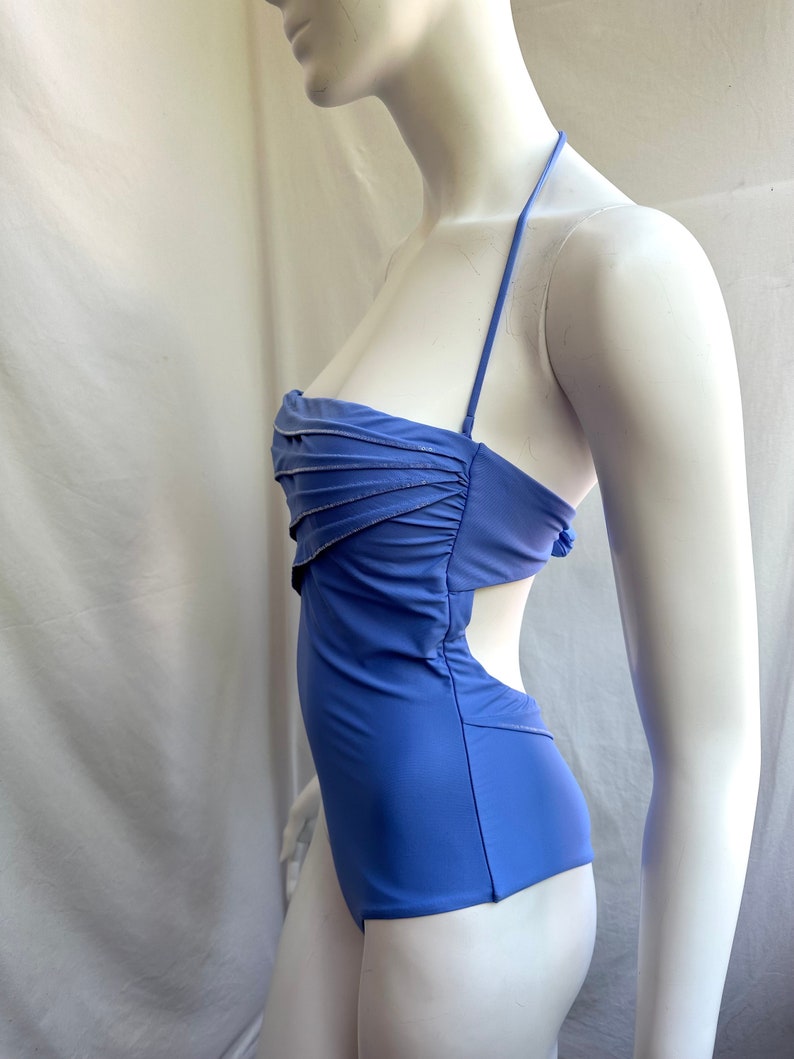 Pu&ograve; includere: Un costume da bagno blu pervinca con collo all'americana e design a taglio. La parte superiore presenta un dettaglio arricciato con un sottile bordo argentato. Il costume intero &egrave; esposto su un manichino.