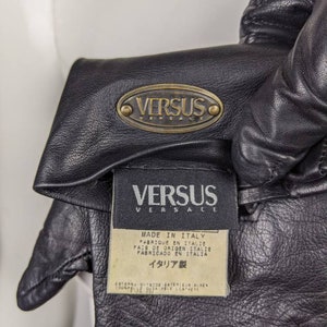 May include: Black leather gloves with a gold oval logo that reads "VERSUS VERSACE" and a white tag that reads "MADE IN ITALY" in multiple languages. The tag also reads "ITALIA製" in Japanese.