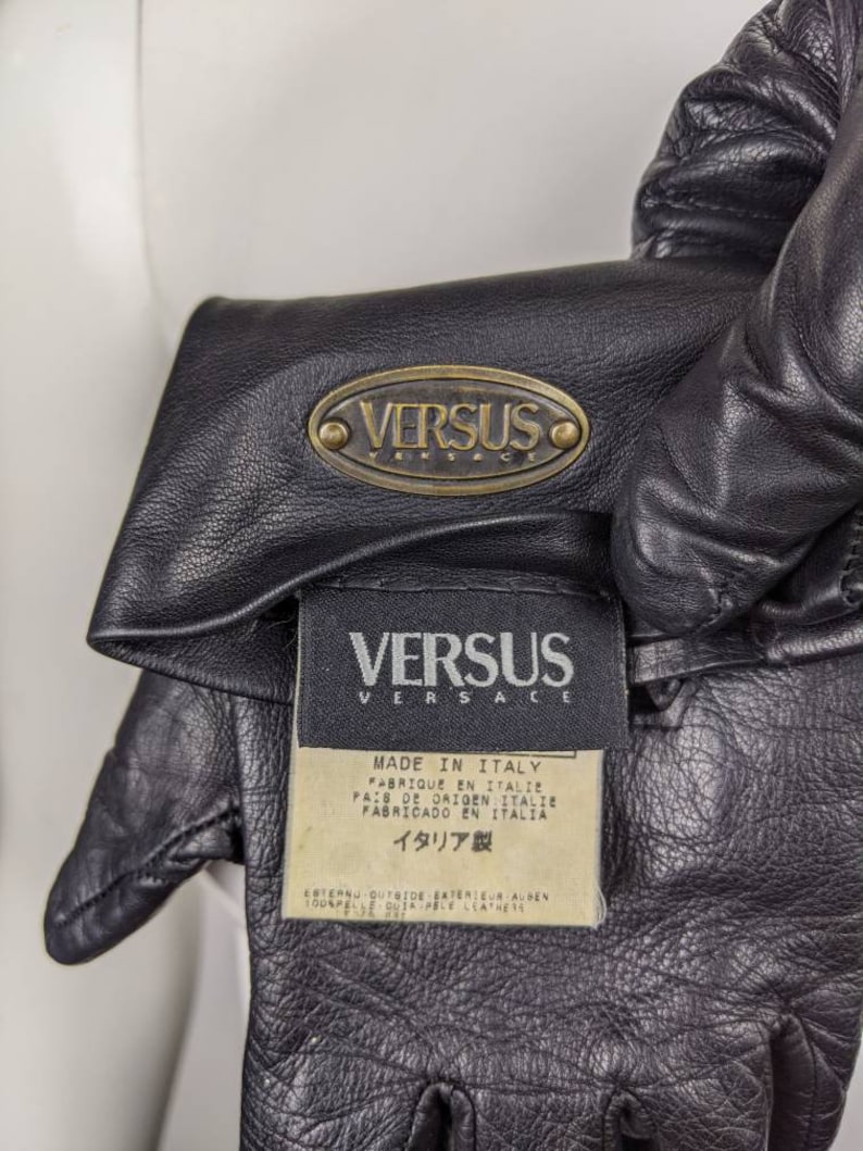 May include: Black leather gloves with a gold oval logo that reads "VERSUS VERSACE" and a white tag that reads "MADE IN ITALY" in multiple languages. The tag also reads "ITALIA製" in Japanese.