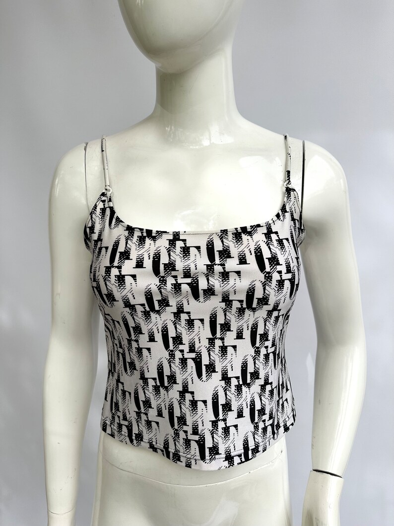 May include: A white tank top with a black and white abstract print. The print features a repeating pattern of geometric shapes and letters.