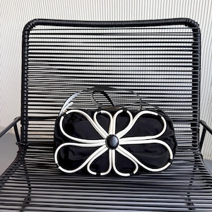 Puede incluir: Un bolso de mano negro con una prominente aplicaci&oacute;n floral blanca en la parte frontal. El dise&ntilde;o consiste en ribetes blancos que forman p&eacute;talos alrededor de un acento circular negro central. El bolso tiene un cuerpo de tela negra lisa.