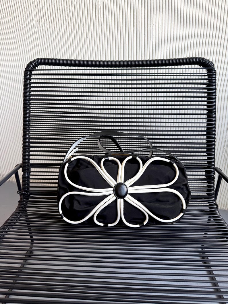 Puede incluir: Un bolso de mano negro con una prominente aplicaci&oacute;n floral blanca en la parte frontal. El dise&ntilde;o consiste en ribetes blancos que forman p&eacute;talos alrededor de un acento circular negro central. El bolso tiene un cuerpo de tela negra lisa.