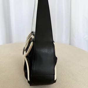 Puede incluir: Un bolso de hombro negro con una silueta estructurada, correa y panel lateral de cuero liso. El exterior est&aacute; acentuado con ribetes blancos en contraste que crean un patr&oacute;n geom&eacute;trico y curvo sobre el cuerpo de tela negra del bolso.