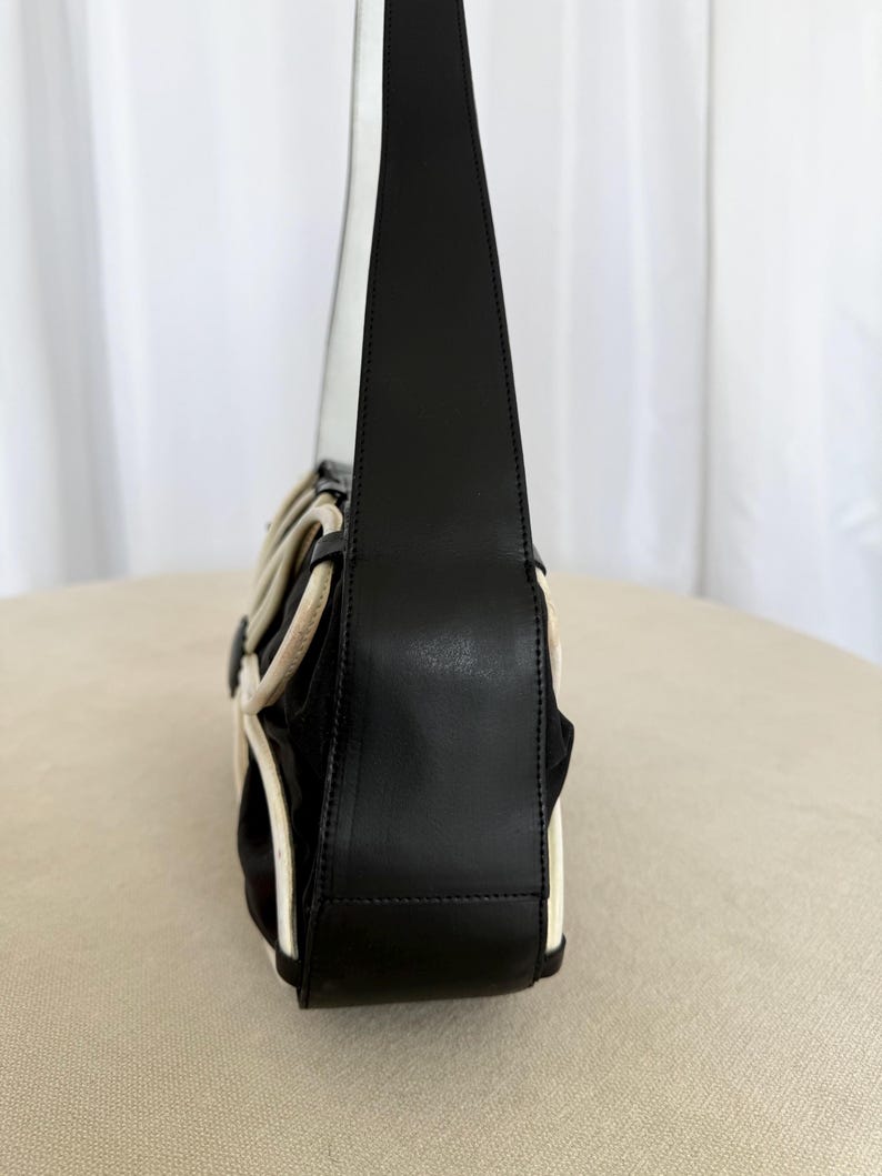 Puede incluir: Un bolso de hombro negro con una silueta estructurada, correa y panel lateral de cuero liso. El exterior est&aacute; acentuado con ribetes blancos en contraste que crean un patr&oacute;n geom&eacute;trico y curvo sobre el cuerpo de tela negra del bolso.