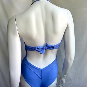Pu&ograve; includere: Un costume da bagno intero blu pervinca con collo all'americana e schiena scoperta. Il costume da bagno presenta un dettaglio a fiocco sul retro e un design a forma di V sul retro. Il tessuto ha una leggera lucentezza.