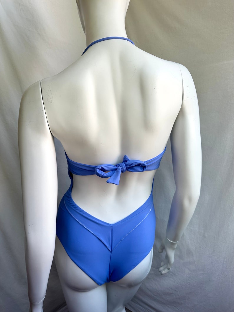 Pu&ograve; includere: Un costume da bagno intero blu pervinca con collo all'americana e schiena scoperta. Il costume da bagno presenta un dettaglio a fiocco sul retro e un design a forma di V sul retro. Il tessuto ha una leggera lucentezza.