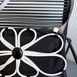 Puede incluir: Un bolso de hombro negro con una gran aplicaci&oacute;n floral blanca en la parte delantera. El dise&ntilde;o de la flor consta de ocho bucles blancos en forma de p&eacute;talo que irradian desde un bot&oacute;n circular negro central. El bolso tiene una correa negra lisa.
