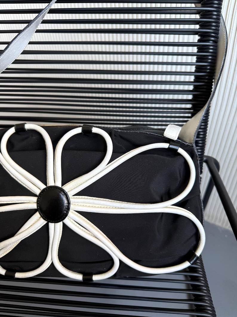 Puede incluir: Un bolso de hombro negro con una gran aplicaci&oacute;n floral blanca en la parte delantera. El dise&ntilde;o de la flor consta de ocho bucles blancos en forma de p&eacute;talo que irradian desde un bot&oacute;n circular negro central. El bolso tiene una correa negra lisa.