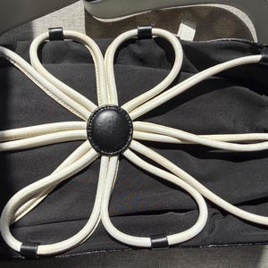 Puede incluir: Un bolso de mano negro con una gran aplicaci&oacute;n floral blanca en la parte frontal. La flor est&aacute; construida con bucles de ribete blanco que irradian desde un bot&oacute;n circular negro central. El bolso tiene un cuerpo de tela negra y una correa blanca.