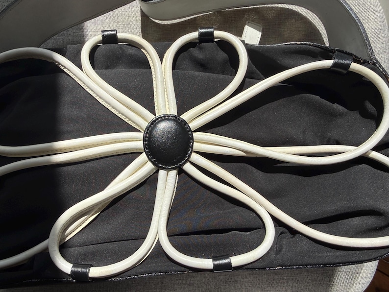 Puede incluir: Un bolso de mano negro con una gran aplicaci&oacute;n floral blanca en la parte frontal. La flor est&aacute; construida con bucles de ribete blanco que irradian desde un bot&oacute;n circular negro central. El bolso tiene un cuerpo de tela negra y una correa blanca.