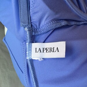 Pu&ograve; includere: Primo piano di un capo color pervinca con un'etichetta bianca con la scritta "LA PERLA" in lettere maiuscole nere. Il tessuto ha una consistenza liscia ed &egrave; cucito con filo azzurro. Il capo sembra essere lingerie.