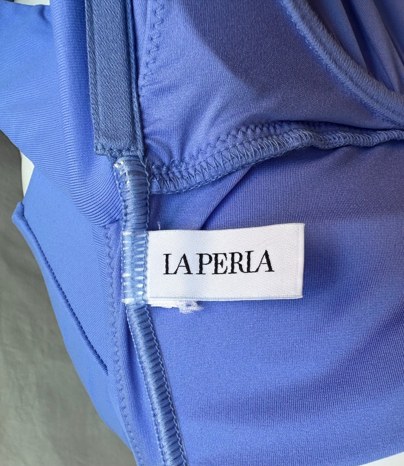Pu&ograve; includere: Primo piano di un capo color pervinca con un'etichetta bianca con la scritta "LA PERLA" in lettere maiuscole nere. Il tessuto ha una consistenza liscia ed &egrave; cucito con filo azzurro. Il capo sembra essere lingerie.