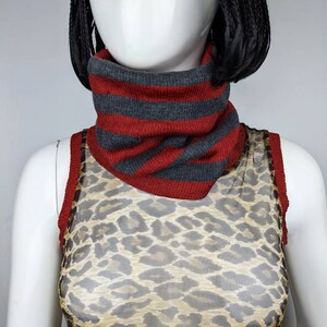 May include: A red and gray striped knit neck warmer. The neck warmer is worn by a mannequin wearing a leopard print mesh top.