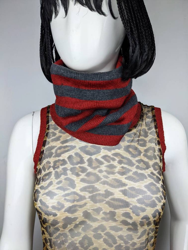 May include: A red and gray striped knit neck warmer. The neck warmer is worn by a mannequin wearing a leopard print mesh top.