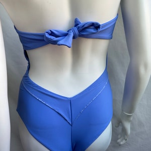 Pu&ograve; includere: Un costume da bagno intero blu pervinca con scollo all'americana e chiusura a fiocco sulla schiena. Il costume da bagno presenta un design a forma di V sul davanti e una schiena scoperta. Il tessuto &egrave; leggermente lucido.