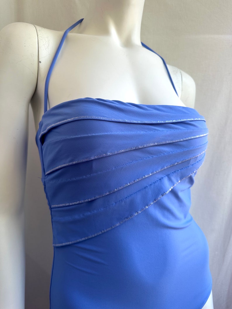 Pu&ograve; includere: Un costume da bagno blu pervinca con collo all'americana e corpetto a strati. Le cuciture argentate accentuano gli strati orizzontali sul busto. Il costume intero &egrave; esposto su un manichino.