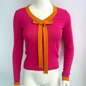 May include: A magenta sweater with long sleeves, an orange V-neck trim, and an orange bow. The cuffs also have orange trim. The sweater is displayed on a mannequin. The bow has long, flowing ties. The sweater is a vibrant piece of clothing.