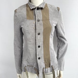 May include: A grey, long-sleeved jacket with vertical stripes and tan panels. The jacket features black buttons, visible stitching, and olive green ties at the bottom. The jacket is displayed on a mannequin.