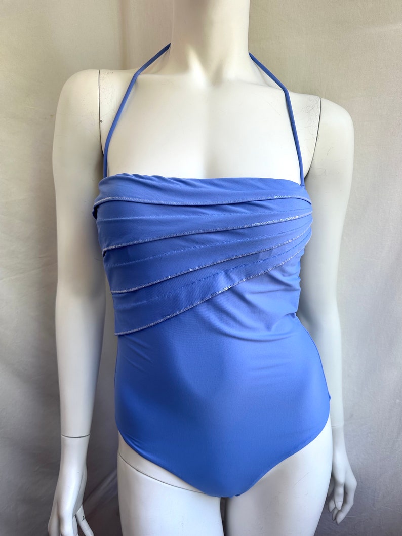 Pu&ograve; includere: Un costume da bagno blu pervinca con collo all'americana e corpetto arricciato. Il costume da bagno presenta accenti argentati lungo le linee arricciate, aggiungendo un tocco di brillantezza. Il design a un pezzo offre un look classico ed elegante.