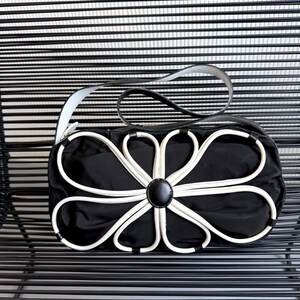 Puede incluir: Un bolso de mano negro con una llamativa aplicaci&oacute;n en forma de flor blanca en la parte frontal. El bolso cuenta con un bot&oacute;n central negro liso, un cuerpo rectangular estructurado y una sola correa de hombro negra con bordes blancos.