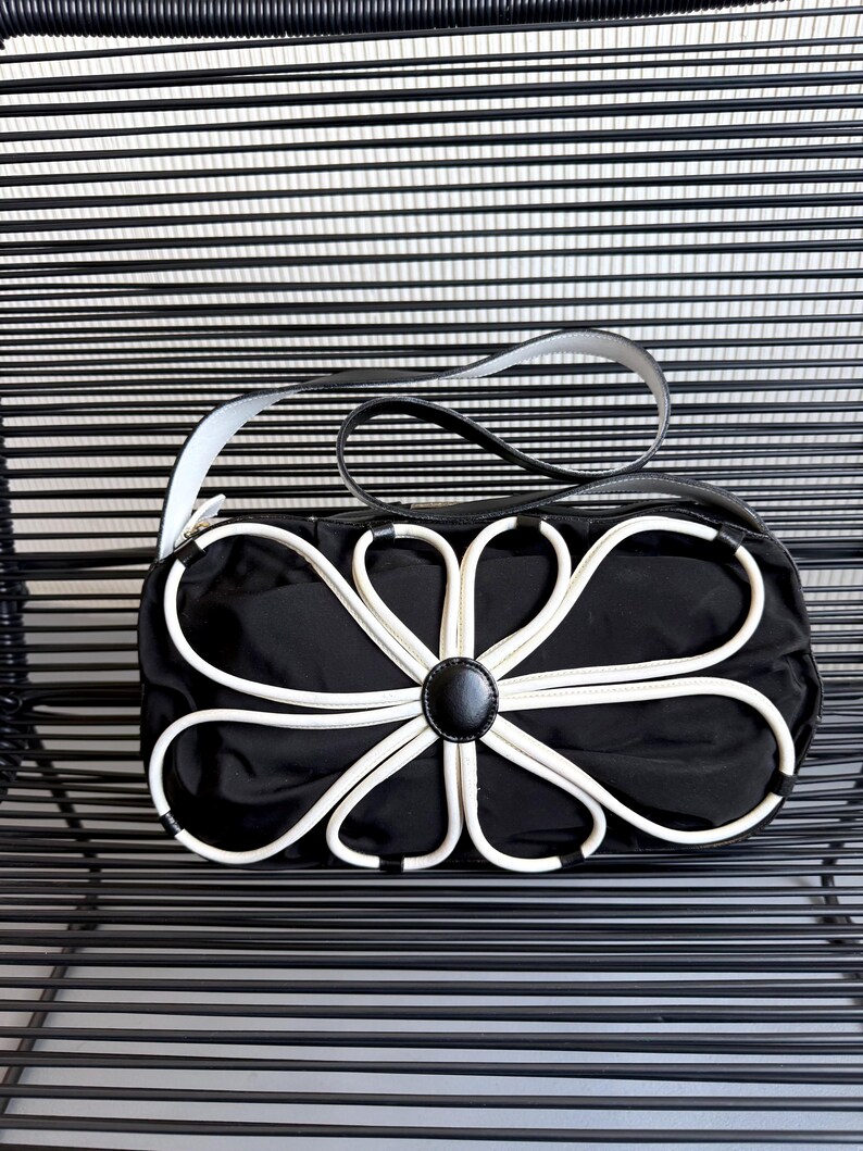 Puede incluir: Un bolso de mano negro con una llamativa aplicaci&oacute;n en forma de flor blanca en la parte frontal. El bolso cuenta con un bot&oacute;n central negro liso, un cuerpo rectangular estructurado y una sola correa de hombro negra con bordes blancos.