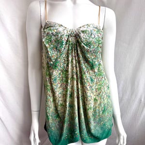 以下が含まれることがあります： 緑、白、青の色合いの花柄のノースリーブトップス。細いゴールドのストラップとバスト部分のノットディテールが特徴です。トップの裾は濃い緑色にグラデーションになっています。