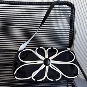 Puede incluir: Un bolso de hombro rectangular negro con una llamativa aplicaci&oacute;n floral blanca en la parte delantera. El dise&ntilde;o floral consiste en ribetes blancos que forman ocho p&eacute;talos alrededor de un bot&oacute;n circular negro central. Incluye una correa larga negra.