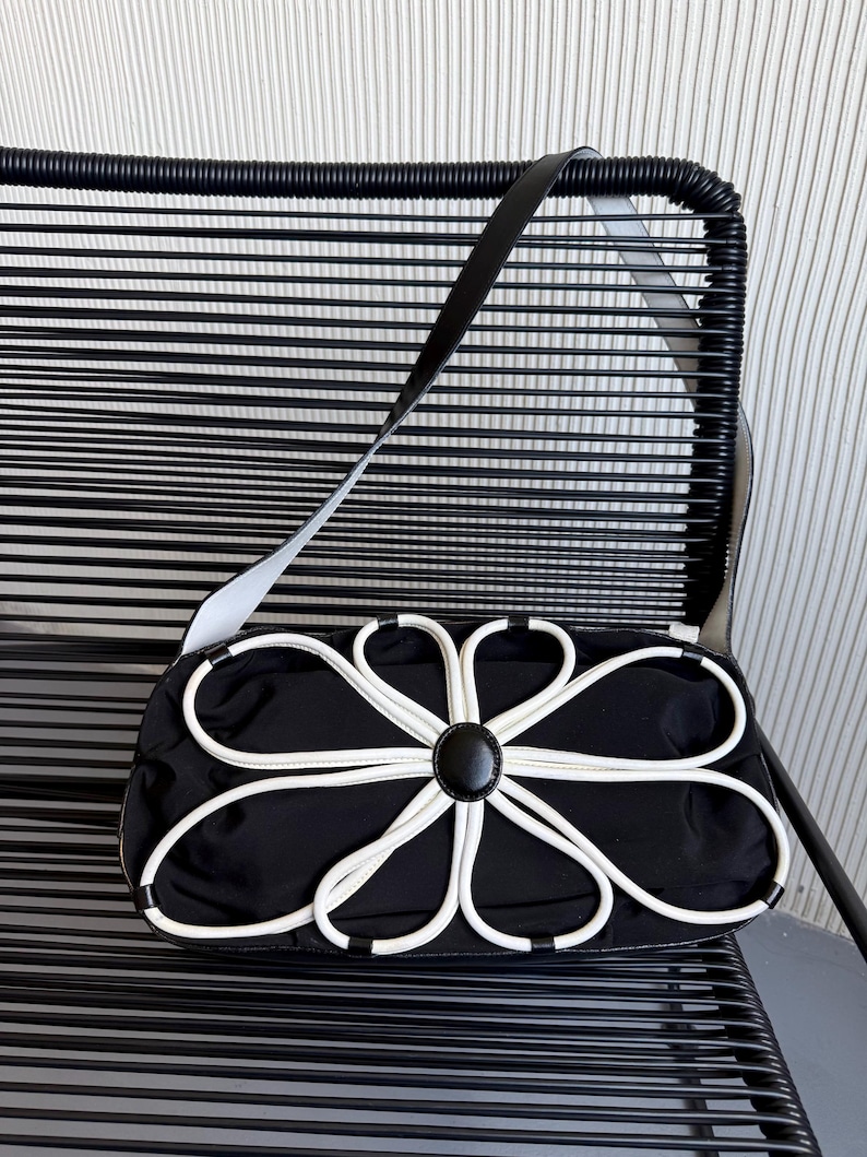 Puede incluir: Un bolso de hombro rectangular negro con una llamativa aplicaci&oacute;n floral blanca en la parte delantera. El dise&ntilde;o floral consiste en ribetes blancos que forman ocho p&eacute;talos alrededor de un bot&oacute;n circular negro central. Incluye una correa larga negra.