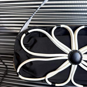 Puede incluir: Un bolso de mano negro con una prominente aplicaci&oacute;n floral blanca en la parte frontal. El dise&ntilde;o de flor se compone de p&eacute;talos de ribete blanco que irradian desde un bot&oacute;n circular negro central. El bolso tiene una correa negra y cuerpo liso.
